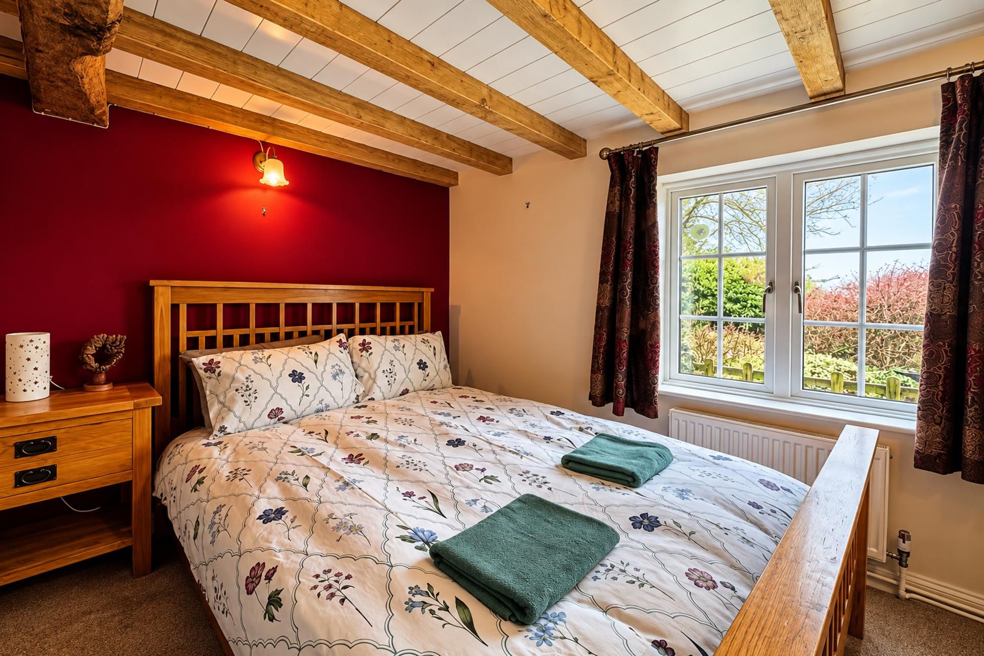 A bedroom with a wooden headboard, floral bedding, a bedside table with a lamp, and a window with curtains.