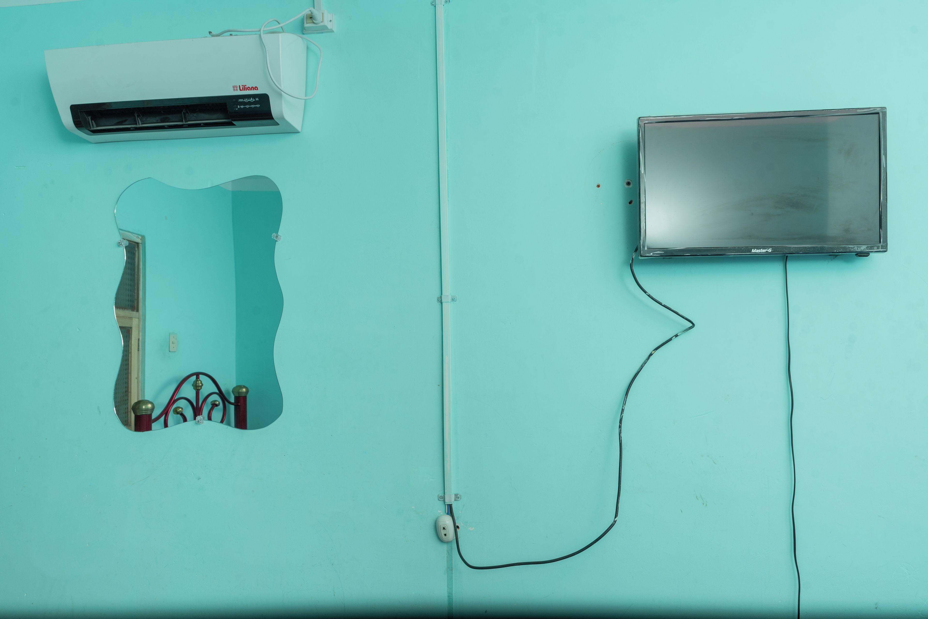 Quarto com parede azul-turquesa, cama com cabeceira vermelha, ar condicionado branco e uma televisão.