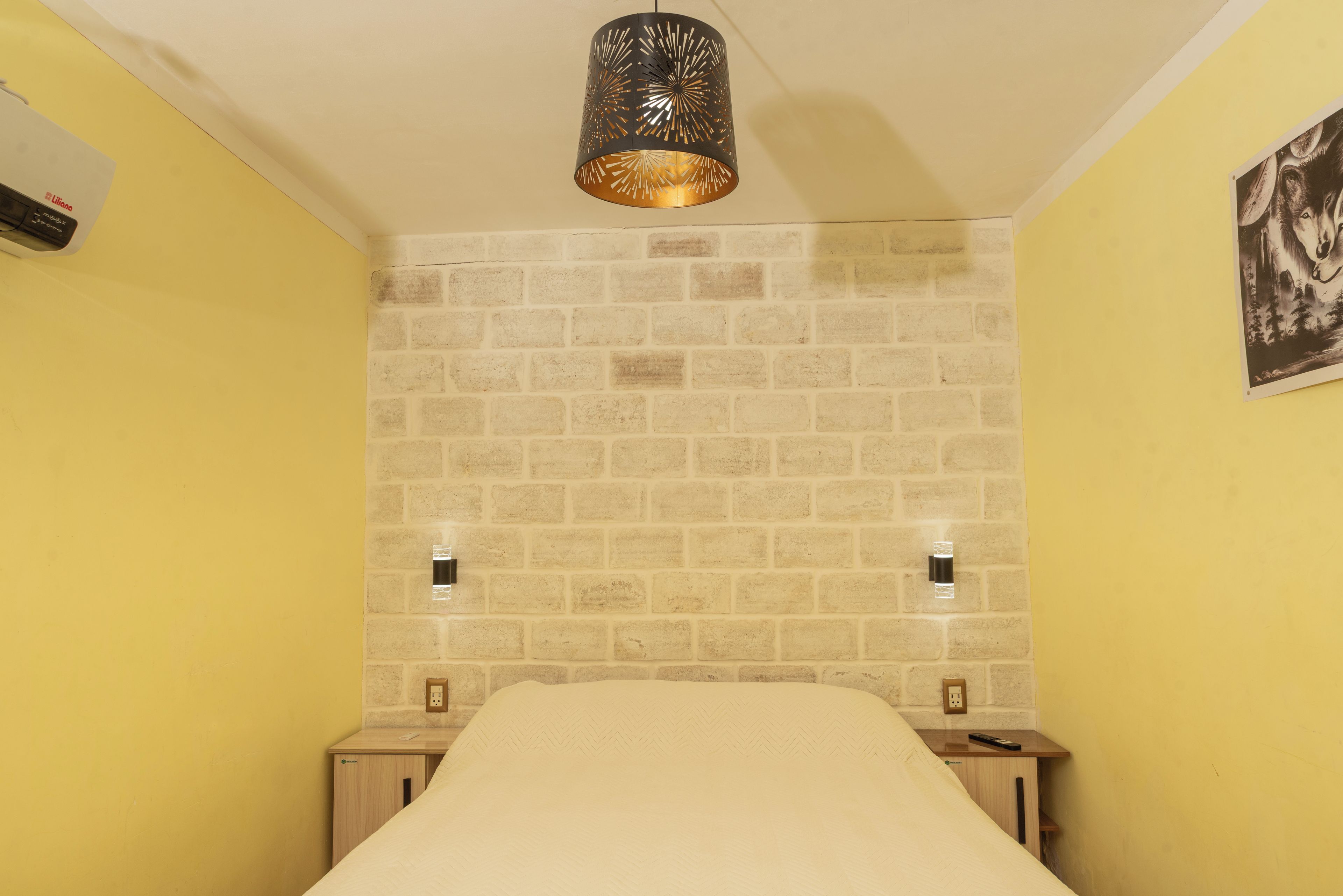 Une pièce avec un mur dont le motif imite un mur de briques, un lit recouvert d’une courtepointe blanche, un luminaire suspendu et une prise de courant fixée au mur.