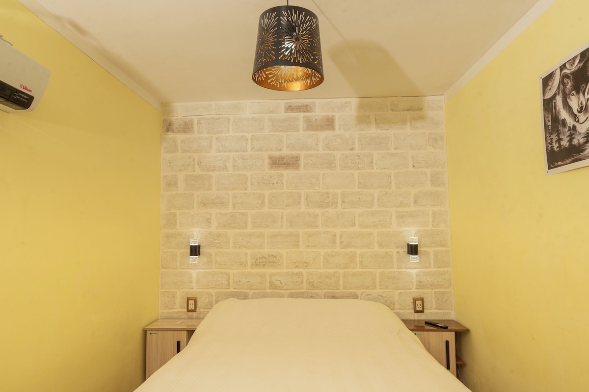 Een kamer met een wand in baksteenmotief, een bed met een witte dekbedovertrek, een hanglamp en een wandcontactdoos.