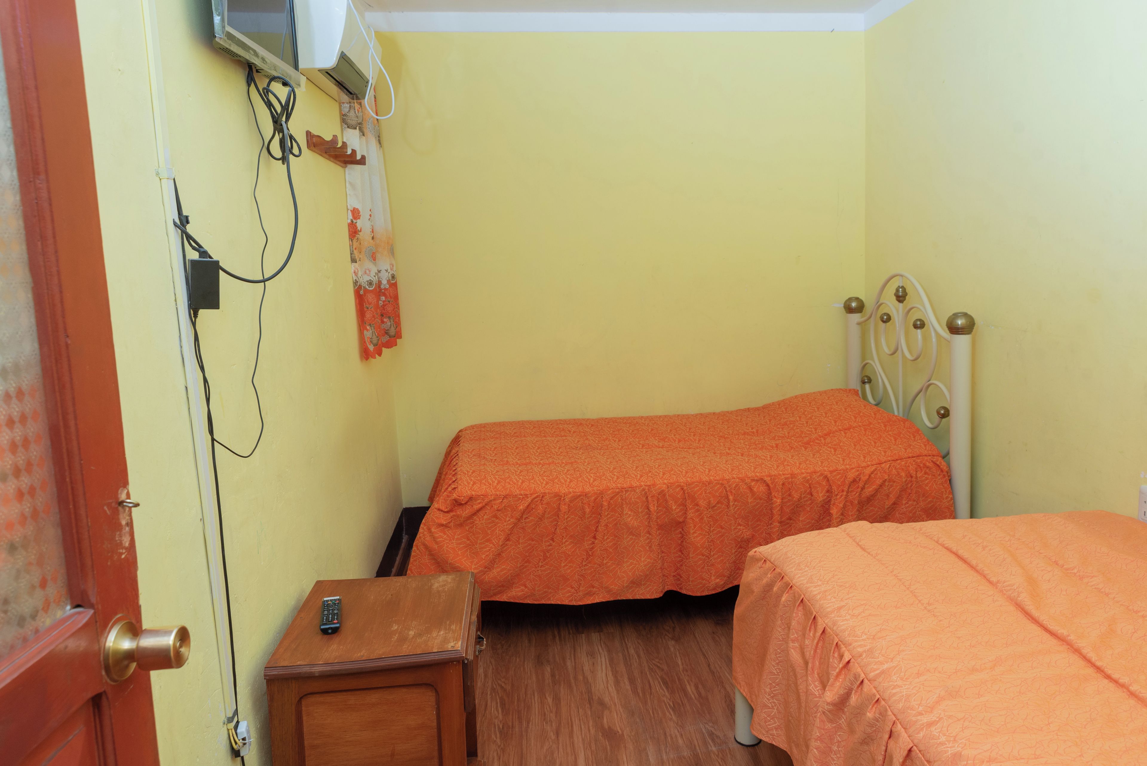 Une petite pièce avec deux lits, une table de chevet en bois, un climatiseur fixé au mur et une porte avec un cadre rouge.