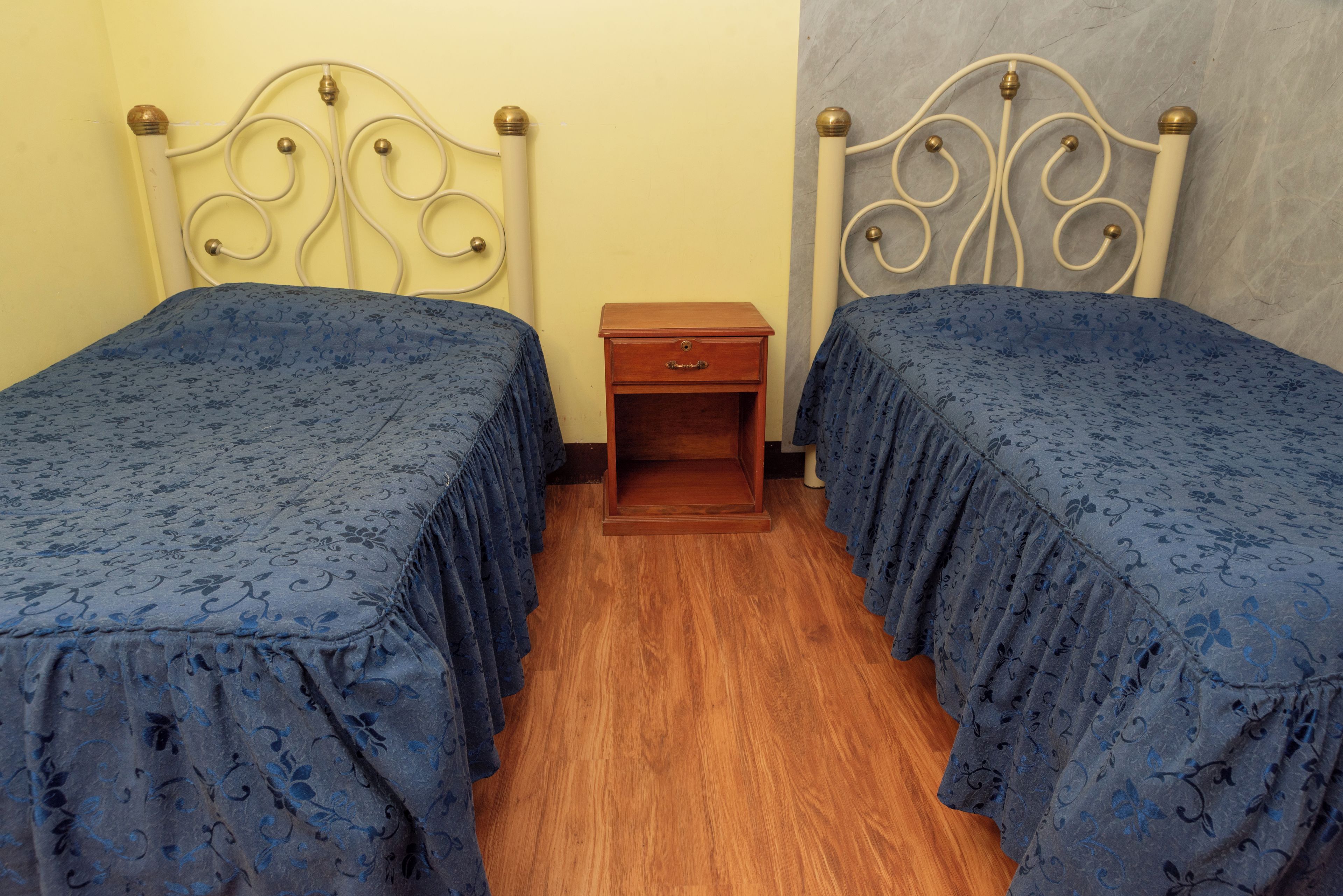 Quarto com duas camas de solteiro, uma mesa de cabeceira de madeira e uma parede lisa.