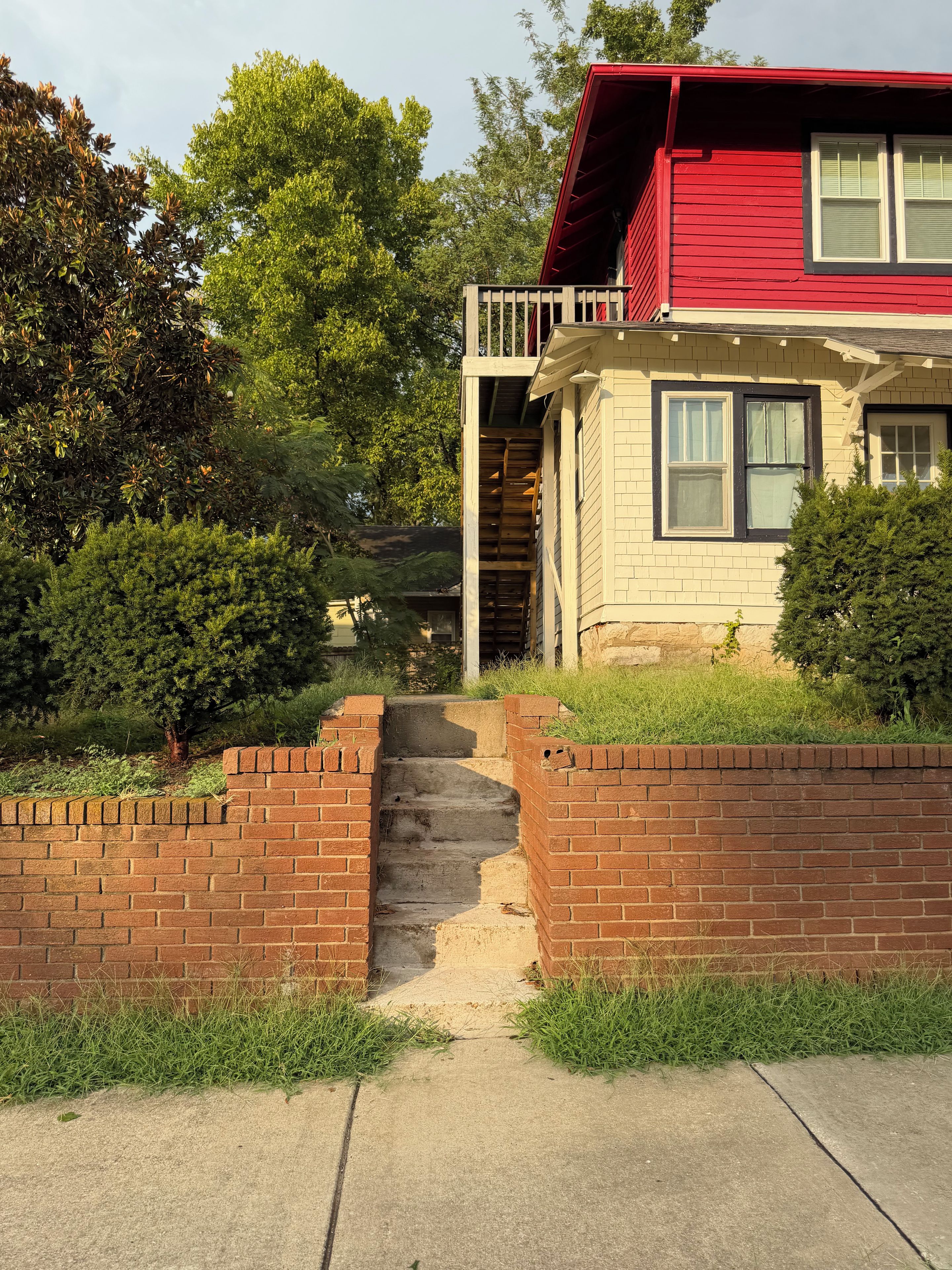 A two-story house with a red roof, a brick staircase leading to a front door, and a small front yard with grass and bushes.