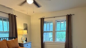 Un dormitorio con cama, ventilador de techo, ventana con cortinas y mesita de noche con lámpara.