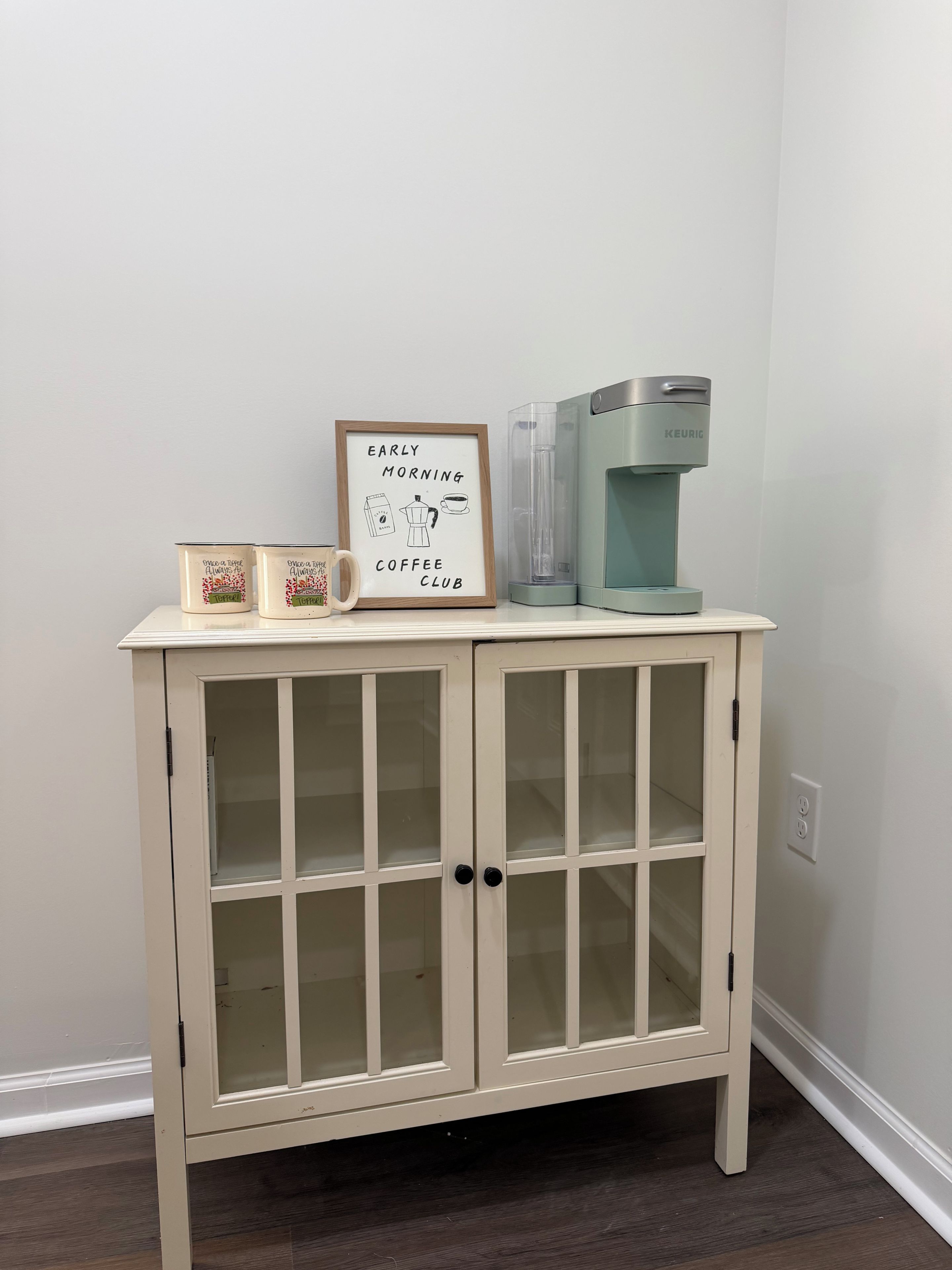 A cabinet with a framed sign, two coffee mugs, and a Keurig coffee maker.