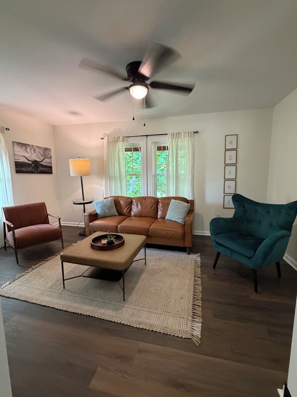 A living room with a brown leather sofa, a teal armchair, a coffee table, a floor lamp, and a ceiling fan.
