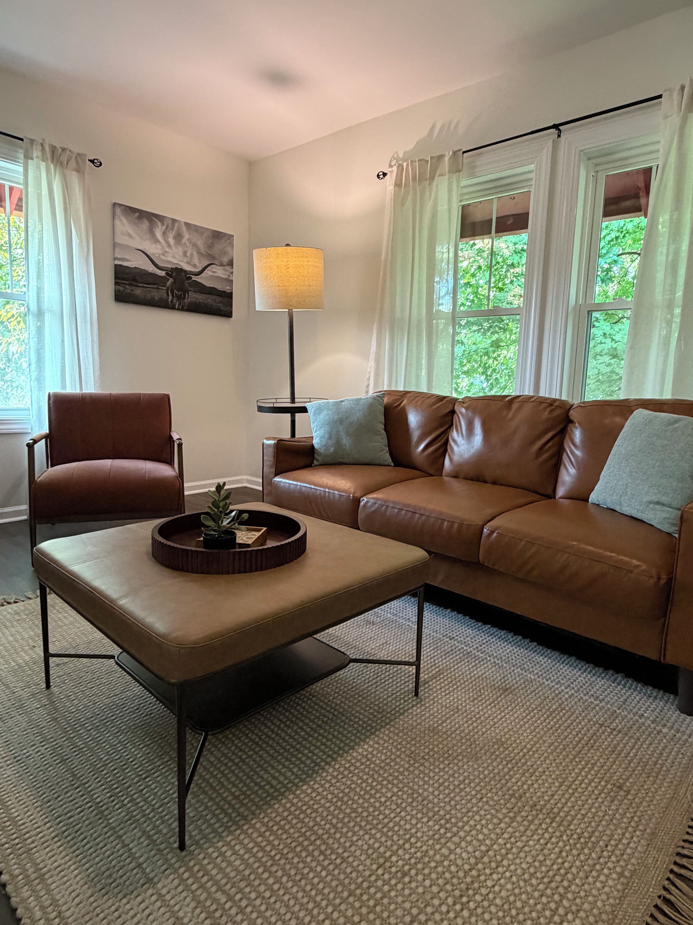 A living room with a brown leather sofa, a brown leather chair, a round coffee table with a plant, a floor lamp, and a painting on the wall.