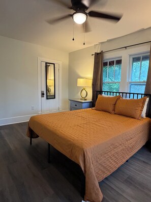 A bedroom with a bed, a nightstand, a lamp, a ceiling fan, a window with curtains, and a door.