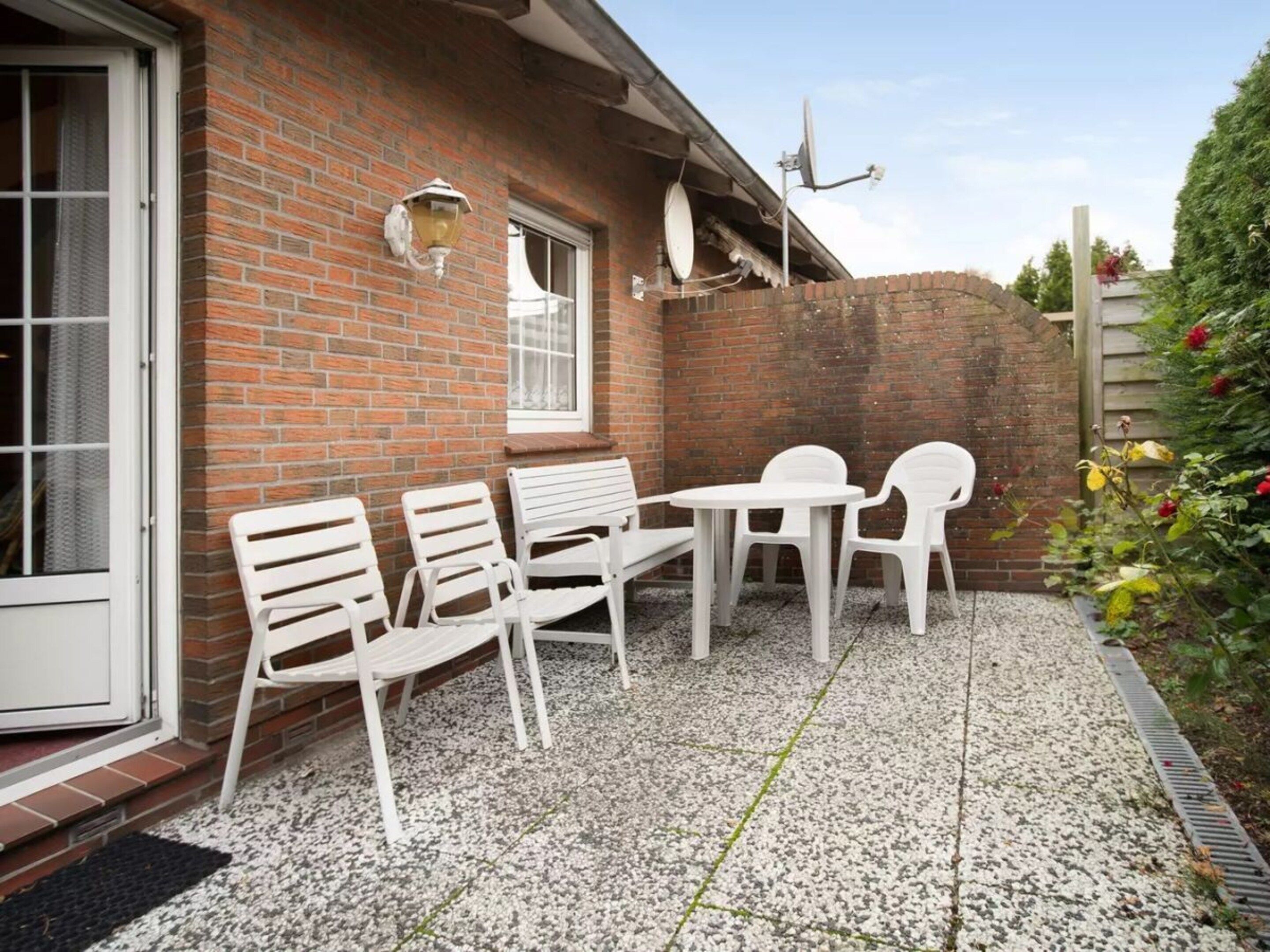 Eine Terrasse mit weißen Plastikstühlen und einem Tisch, einer Ziegelwand und einer Satellitenschüssel.