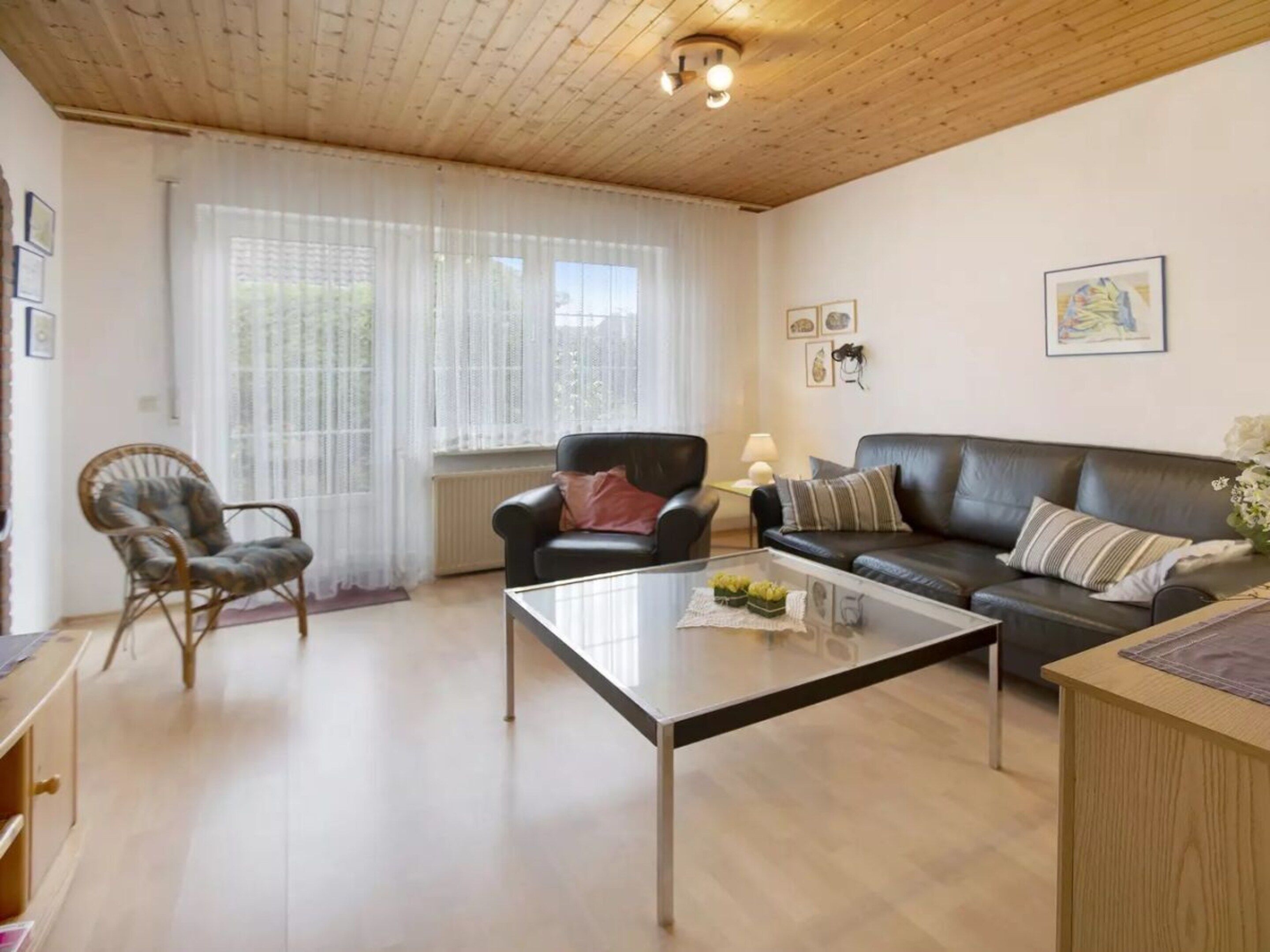 Een woonkamer met een houten plafond, een glazen salontafel, een zwarte leren bank, een rieten fauteuil en een radiator.