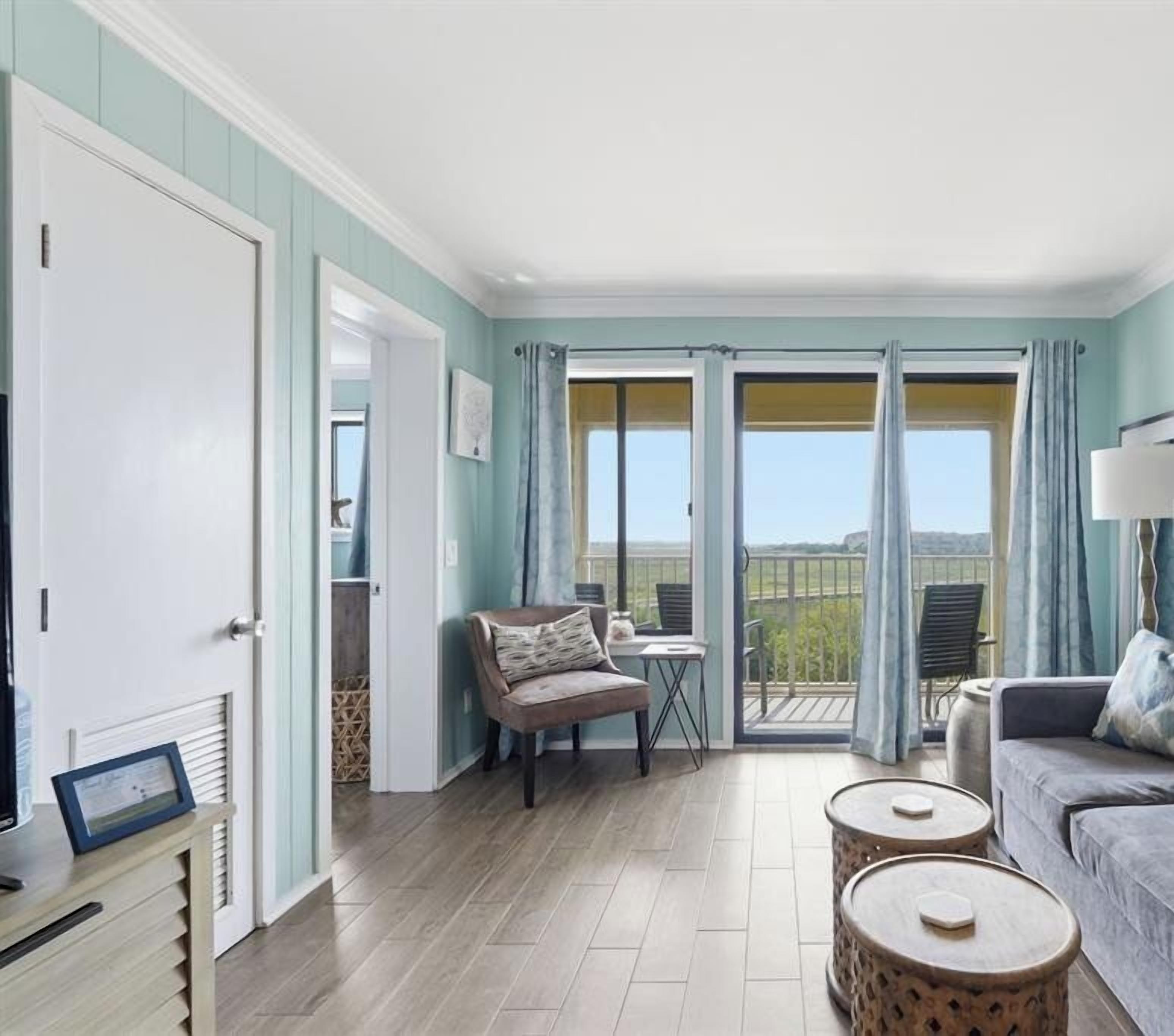 Aqua walls and crisp white trim frame a sun-drenched living area where plush textures meet smooth wood floors. Large glass doors pull the outdoors in, creating a bright sanctuary for quiet afternoons by the balcony.