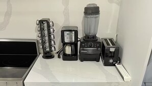 Uma cozinha com bancada, cafeteira, liquidificador e torradeira. Há também algumas canecas e uma panela nas gavetas abaixo.