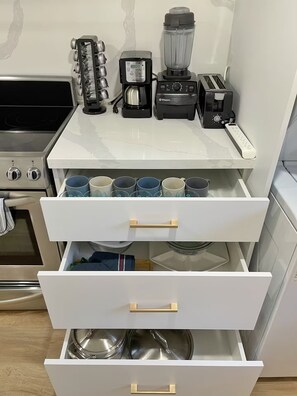 Et køkken med bordplade, kaffemaskine, blender og brødrister. Der er også flere krus og en gryde i skufferne nedenunder.