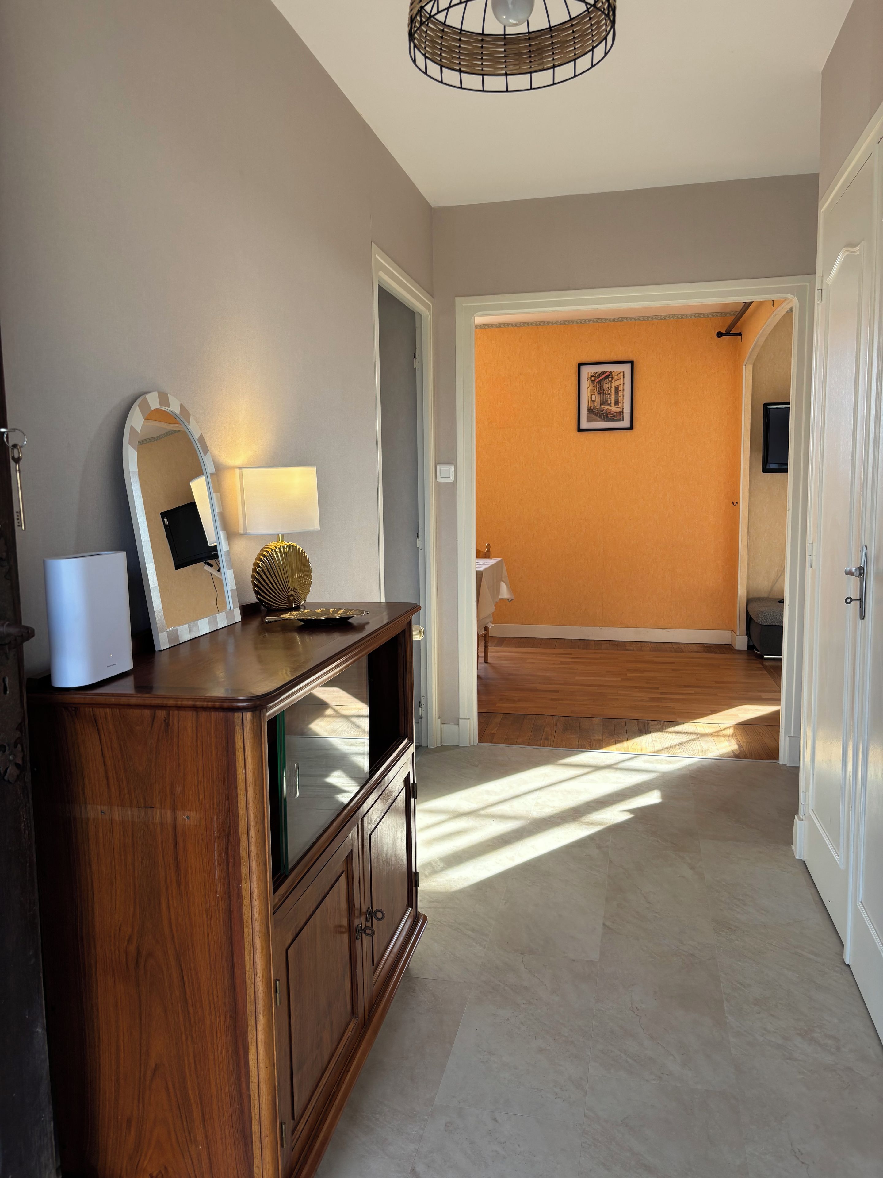 Um corredor com um armário de madeira, um espelho e uma luminária. Há uma porta que dá para outro quarto com um quadro na parede.