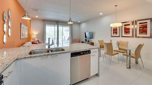 Cozinha moderna com ilha central, área de refeições com mesa de vidro e sala de estar com TV de tela plana.