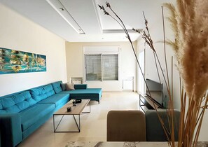 A modern living room with a sectional sofa, a coffee table, a flat-screen TV, and a large window with blinds. - Perfect Landing Pad (Gouvernorat de l'Ariana)
