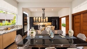 A modern kitchen with dark cabinetry, a marble countertop, and a dining area with a glass table and chairs. - Villa Punta Espada CapCana, 6-bedroom, with pool and hole 8 (Punta Cana)
