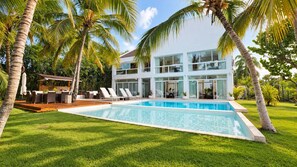Uma casa moderna de dois andares com piscina, cercada por palmeiras e um gramado bem cuidado.