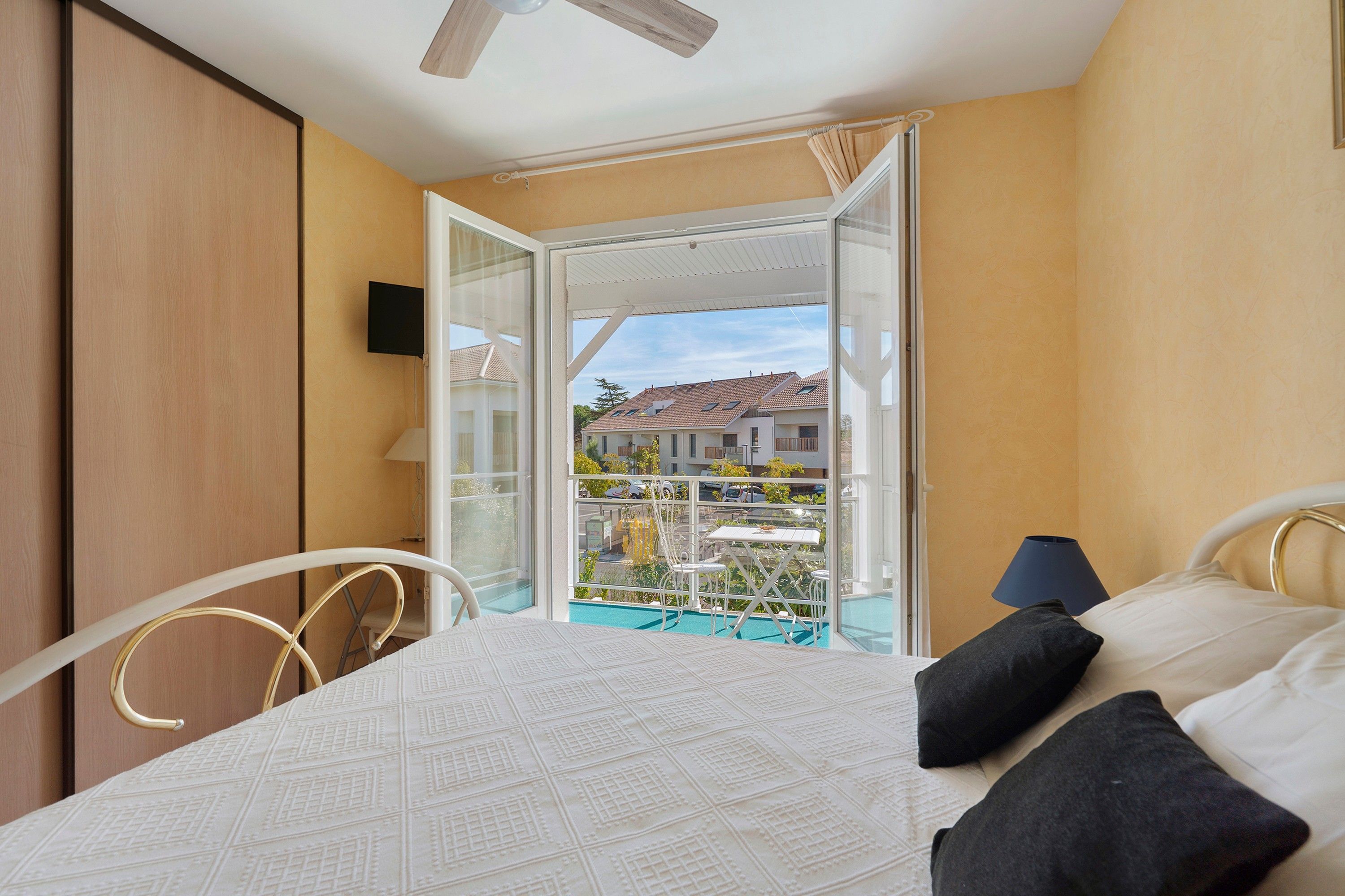 Une chambre à coucher avec un lit, un balcon donnant sur un immeuble et un ventilateur de plafond.