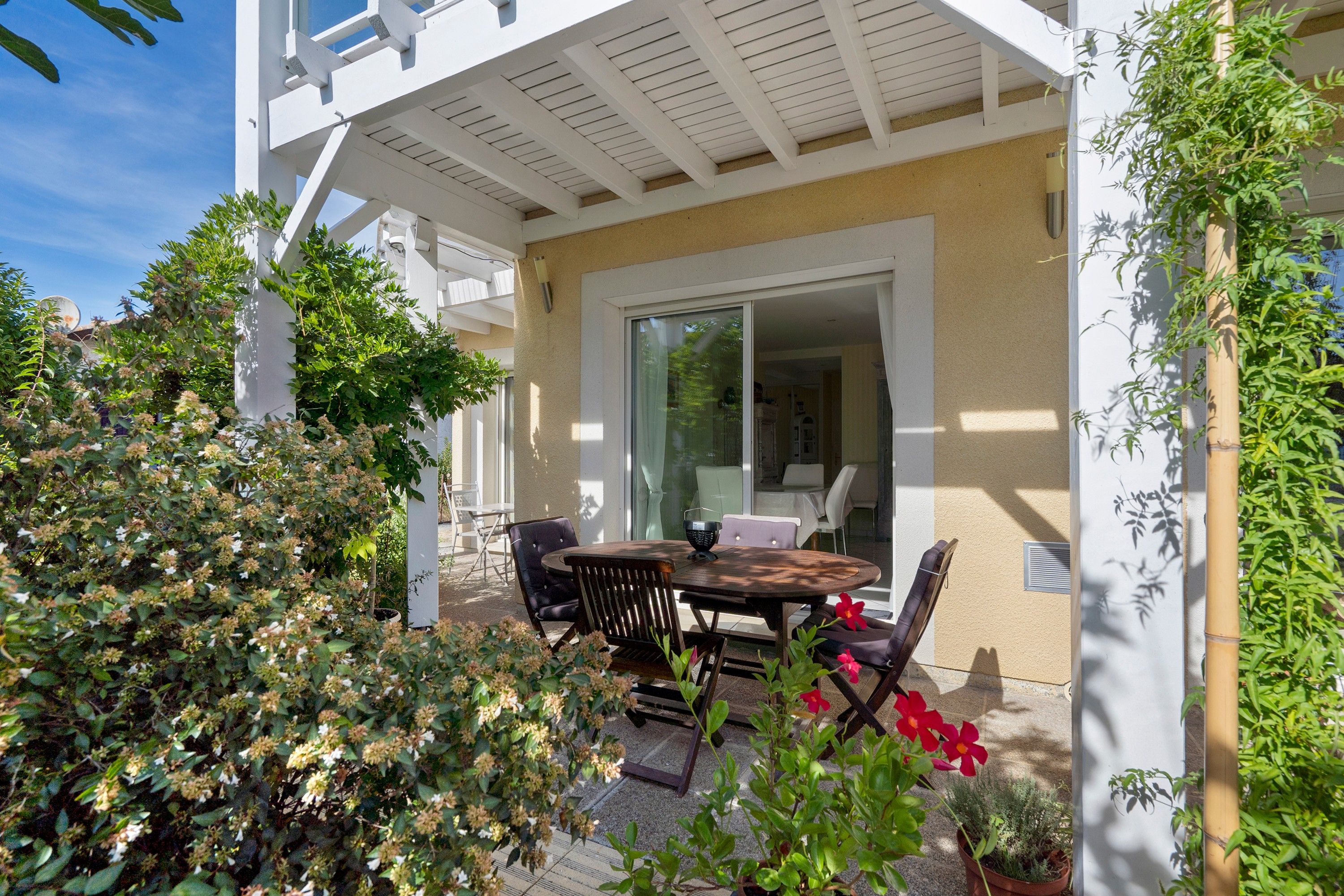 Une terrasse avec une table et des chaises, entourée de verdure et bordée d’un bâtiment à la façade blanche.