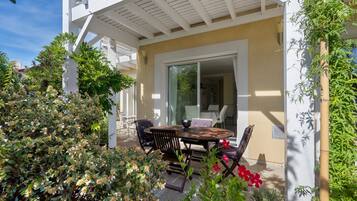 Une terrasse avec une table et des chaises, entourée de verdure et bordée d’un bâtiment à la façade blanche.