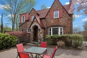 A brick house with a stone archway entrance, a patio with a table and chairs, and a red flowering tree.