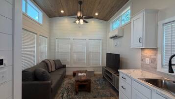 A compact living space with a ceiling fan, a sofa, a coffee table, a TV, a kitchen area with cabinets and a sink, and a window with blinds.