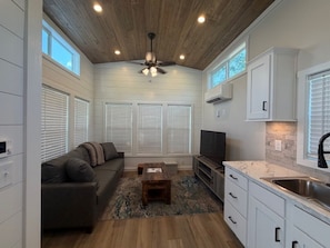 A compact living space with a ceiling fan, a sofa, a coffee table, a TV, a kitchen area with cabinets and a sink, and a window with blinds. - Magnolia MS Riverbend (Vicksburg)