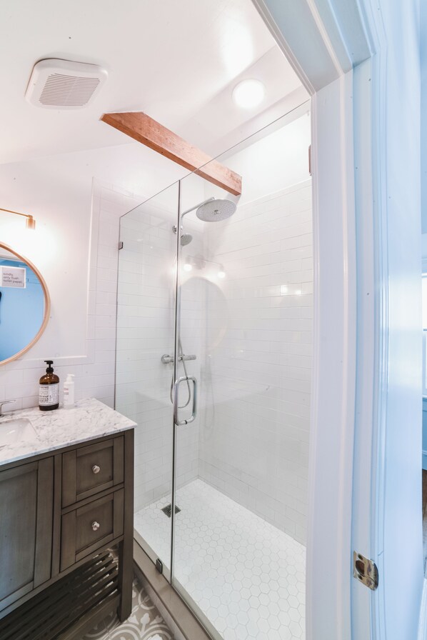 A modern bathroom with a glass-enclosed shower, white tiled walls, a marble countertop, and a wooden shelf above the sink.