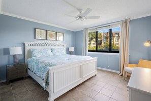 A bedroom with a bed, nightstands, a ceiling fan, a window with curtains, and a view of buildings outside. - Oceanfront Retreat Patio To Beach Pool Hot Tub (Hilton Head Island)