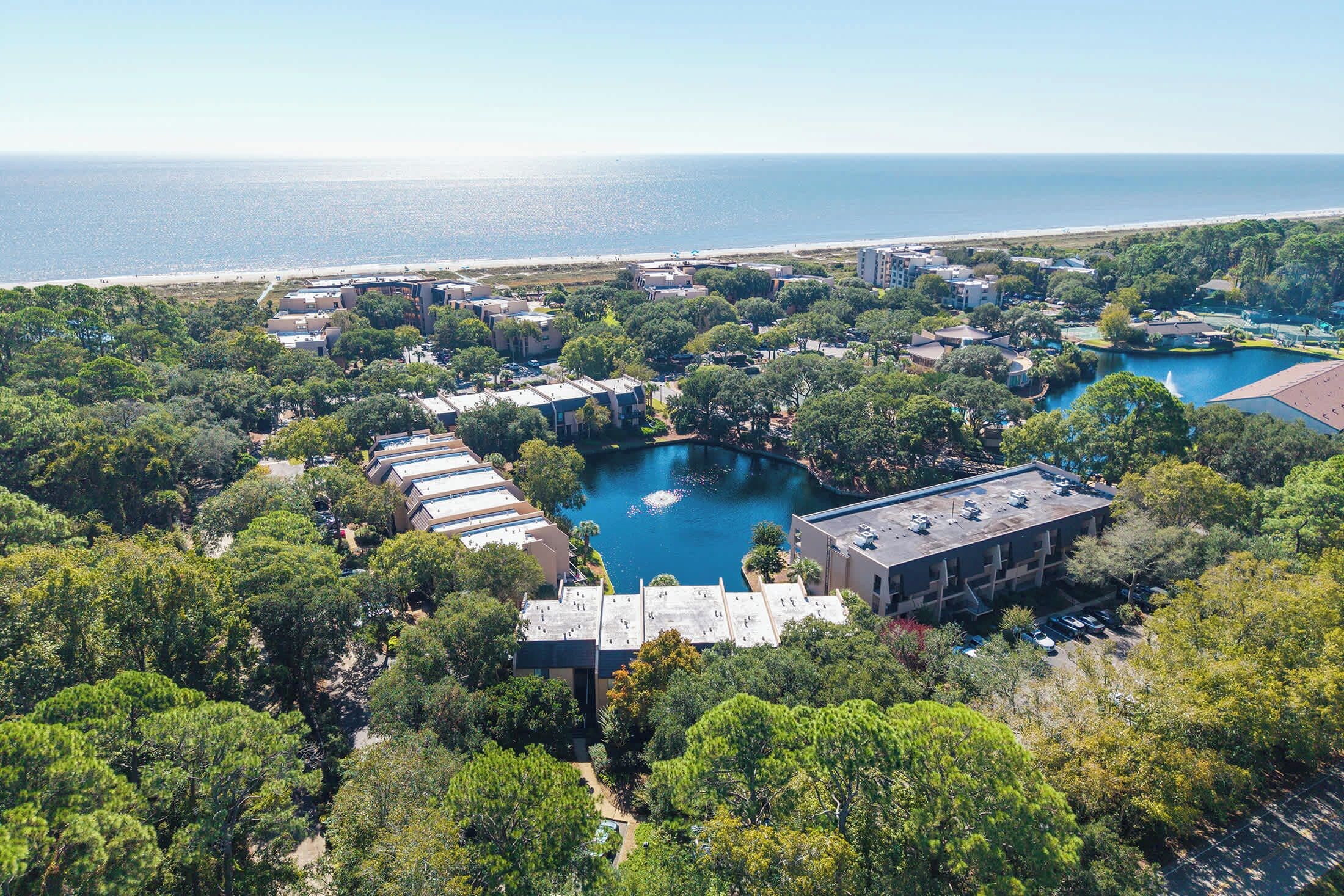 An aerial view of a residential area with multiple buildings, a central water feature, and a beach in the background.