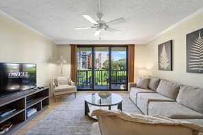 A living room with a flat-screen TV, a ceiling fan, a sofa, a coffee table, and a view of a balcony with outdoor furniture. - Oceanfront Retreat Patio To Beach Pool Hot Tub (Hilton Head Island)