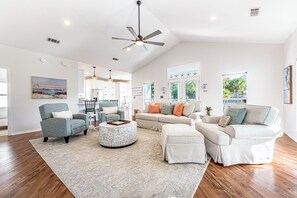 A spacious living room with a high ceiling, a ceiling fan, a dining area with a kitchen in the background, and large windows providing natural light. - Beach Cottage with Golf Cart & Community Pool (Destin)
