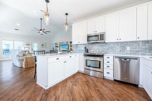 A modern kitchen with white cabinetry, a central island, stainless steel appliances, and a view of the outdoors. - Beach Cottage with Golf Cart & Community Pool (Destin)