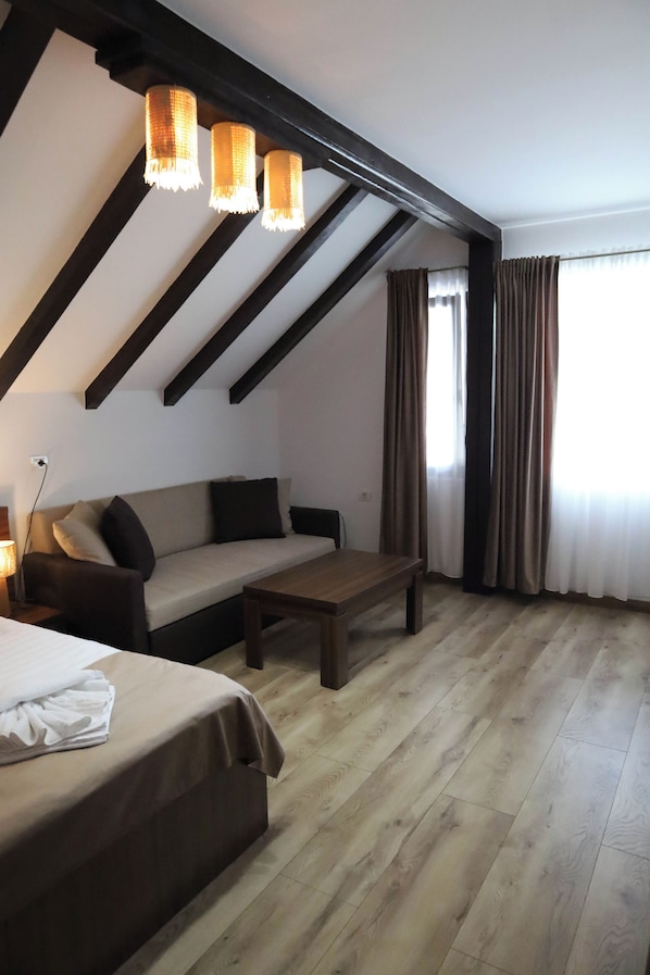 A room with a sofa, a wooden coffee table, a bed with a white cover, and a window with curtains.
