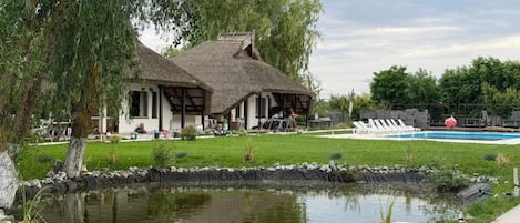 Una casa con techo de paja, un estanque y una piscina al fondo.