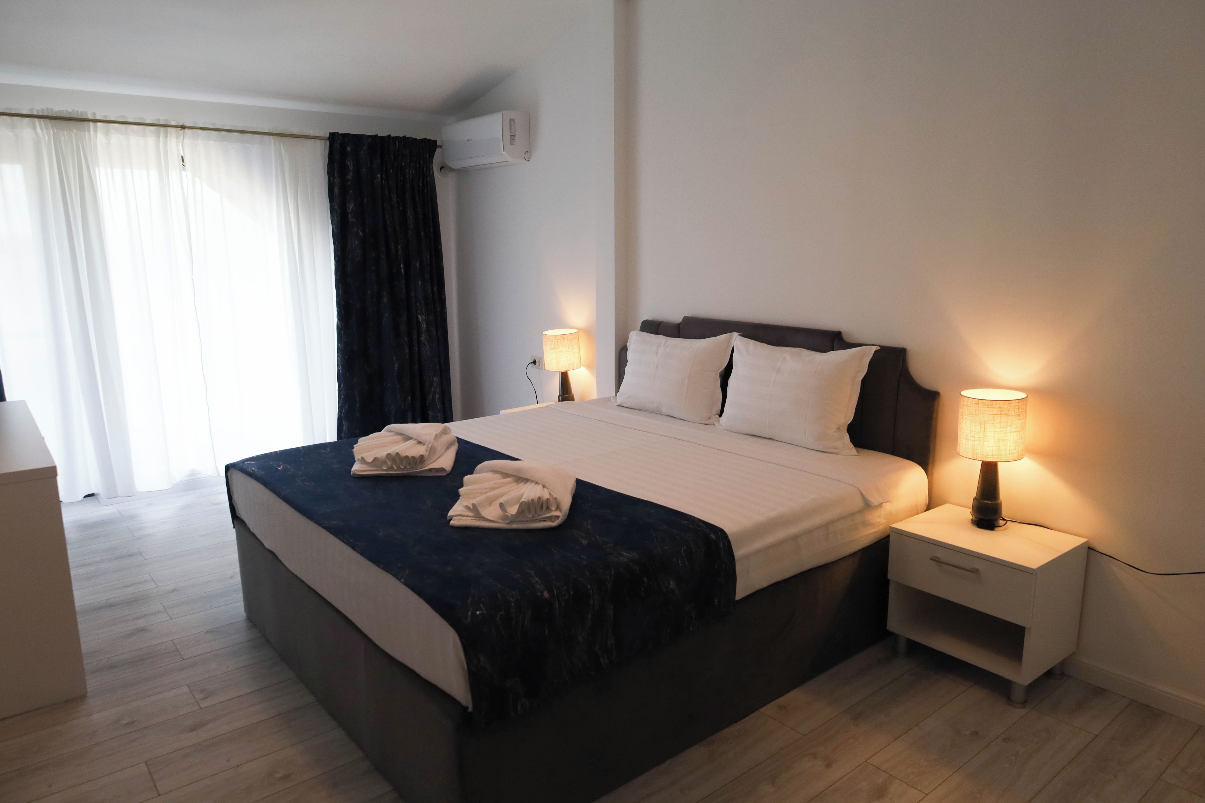 A hotel room with a bed, two bedside lamps, a nightstand, a window with curtains, and an air conditioning unit.
