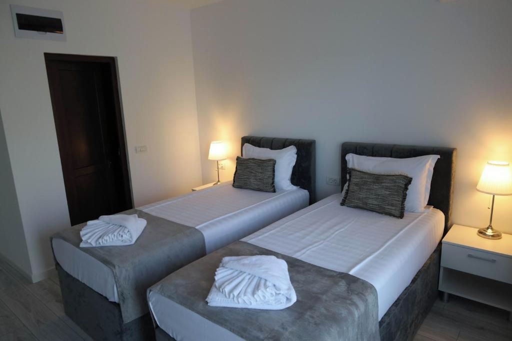 Habitación de hotel con dos camas, cada una con una almohada y una toalla doblada. Mesitas de noche con lámpara a cada lado de las camas.