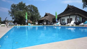 Una piscina con sillas de descanso y sombrillas, un edificio con techo de paja y árboles al fondo.