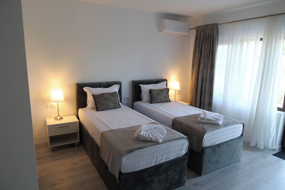 A hotel room with two beds, bedside lamps, an air conditioner, and a window with curtains.