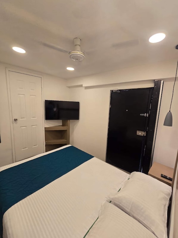Quarto com cama, televisão na parede, ventilador de teto e porta.