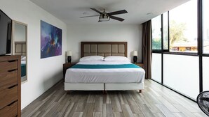 Une chambre d’hôtel moderne avec un grand lit, un ventilateur de plafond, un tableau au mur et une vue sur l’extérieur grâce à de grandes fenêtres.