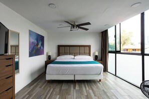 Quarto de hotel moderno com cama grande, ventilador de teto, pintura na parede e vista para o exterior através de grandes janelas.