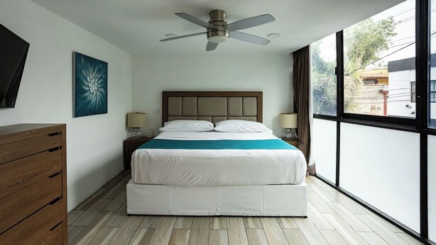 Une chambre moderne avec un lit, des lampes de chevet, un ventilateur de plafond, une commode en bois et une grande fenêtre donnant sur l’extérieur.