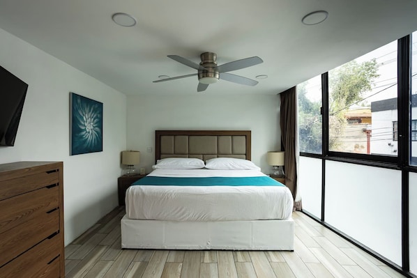 Quarto moderno com cama, abajures, ventilador de teto, cômoda de madeira e uma grande janela com vista para o exterior.