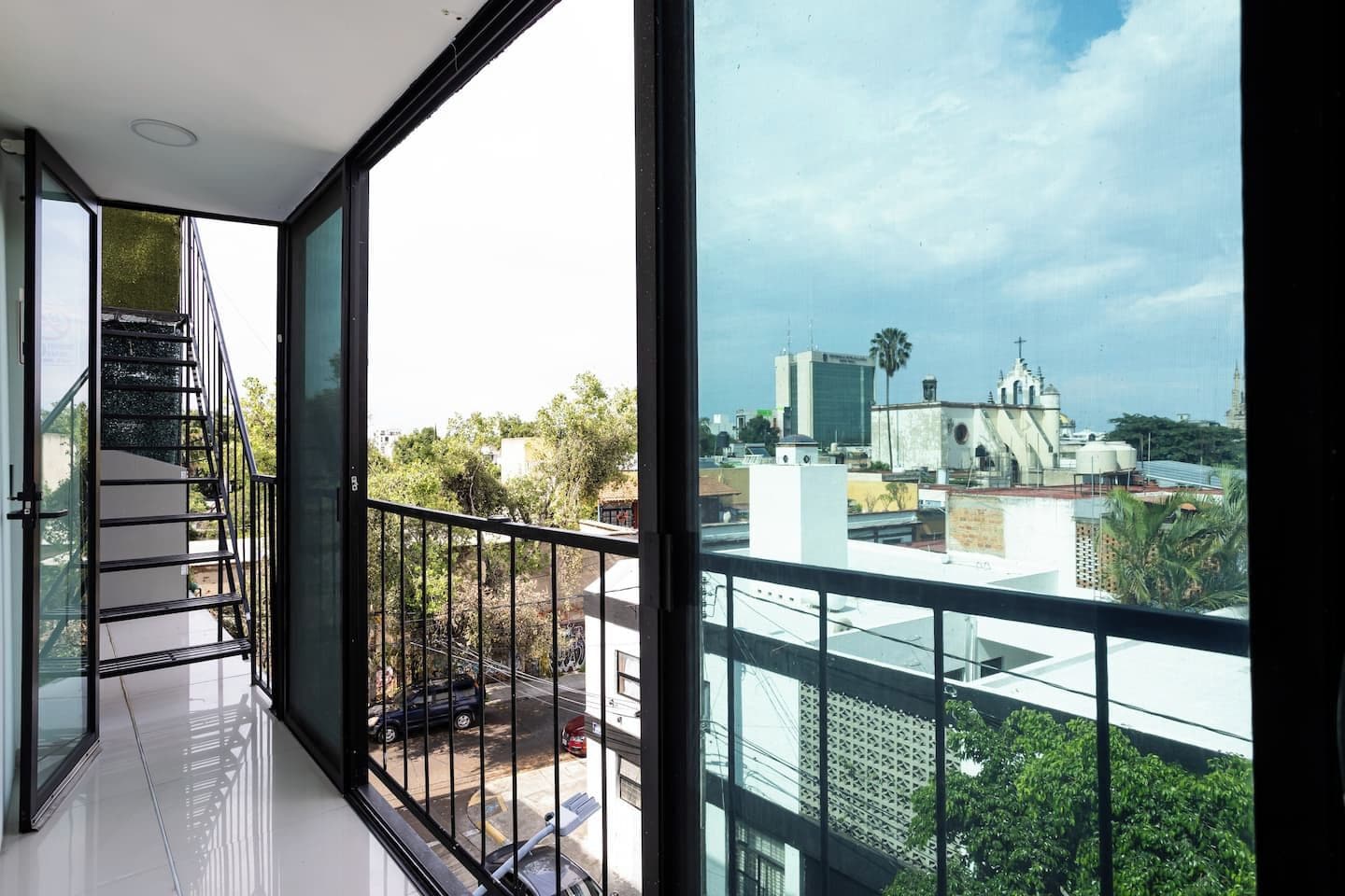 A balcony with a glass door, a metal railing, and a view of a cityscape including buildings and trees.
