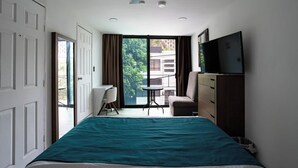 Une chambre à coucher comprenant un lit, un bureau, une chaise, un téléviseur et un balcon offrant une vue sur les bâtiments et les arbres.