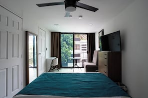 Quarto com cama, escrivaninha, cadeira, televisão e varanda com vista para prédios e árvores.