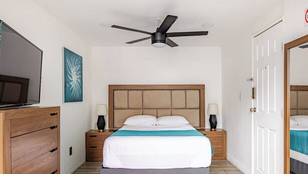 Une chambre d’hôtel avec un lit, des lampes de chevet, un ventilateur de plafond, une télévision et une commode en bois.
