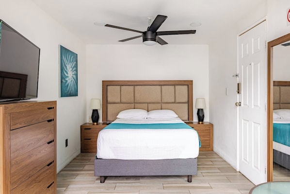 Quarto de hotel com cama, luminárias de cabeceira, ventilador de teto, televisão e cômoda de madeira.